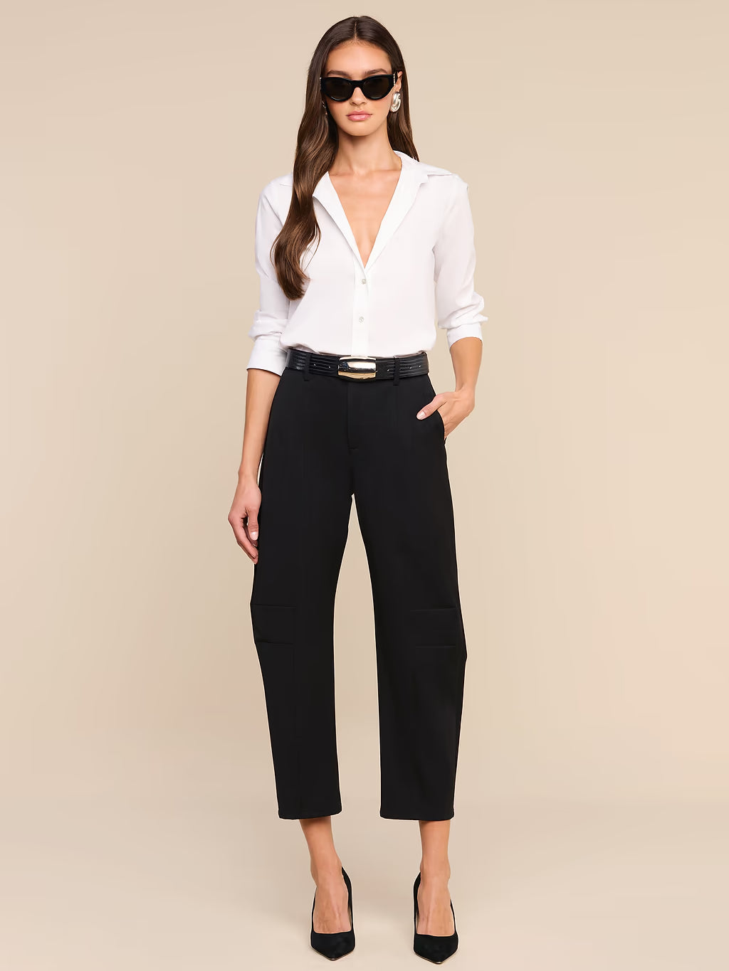 A woman modeling a white button-down shirt tucked into black cropped trousers with a black belt, styled with black heels and sunglasses against a solid beige background.