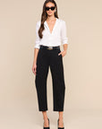 A woman modeling a white button-down shirt tucked into black cropped trousers with a black belt, styled with black heels and sunglasses against a solid beige background.