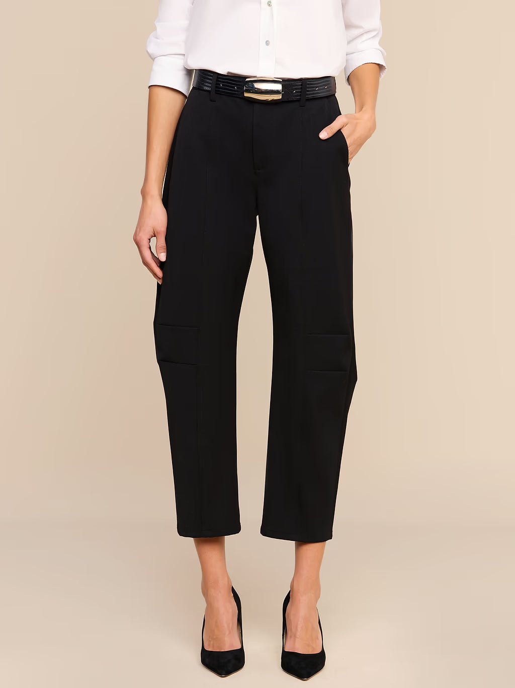 A model wearing black cropped trousers with side pockets, styled with a black belt, a white button-down shirt, and black heels against a solid beige background.