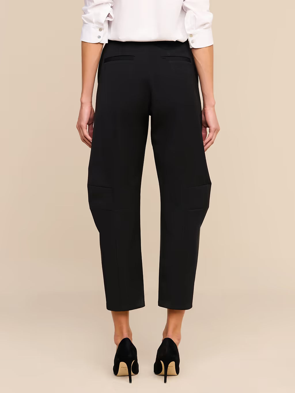 Rear view of a model wearing black cropped trousers with side pockets, paired with a white blouse and black heels, against a solid beige background.