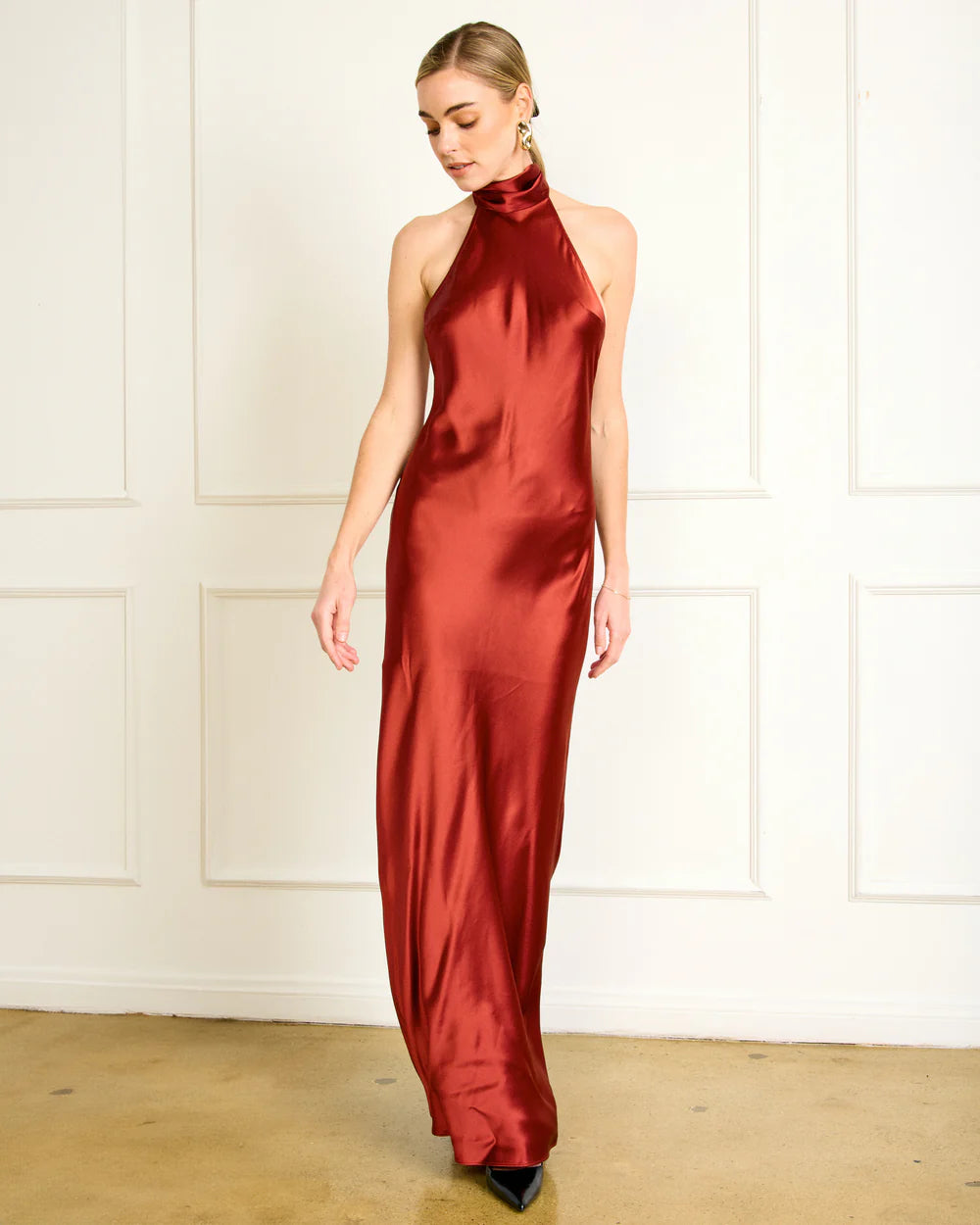 Woman wearing a red satin halter neck gown against a white paneled wall, front