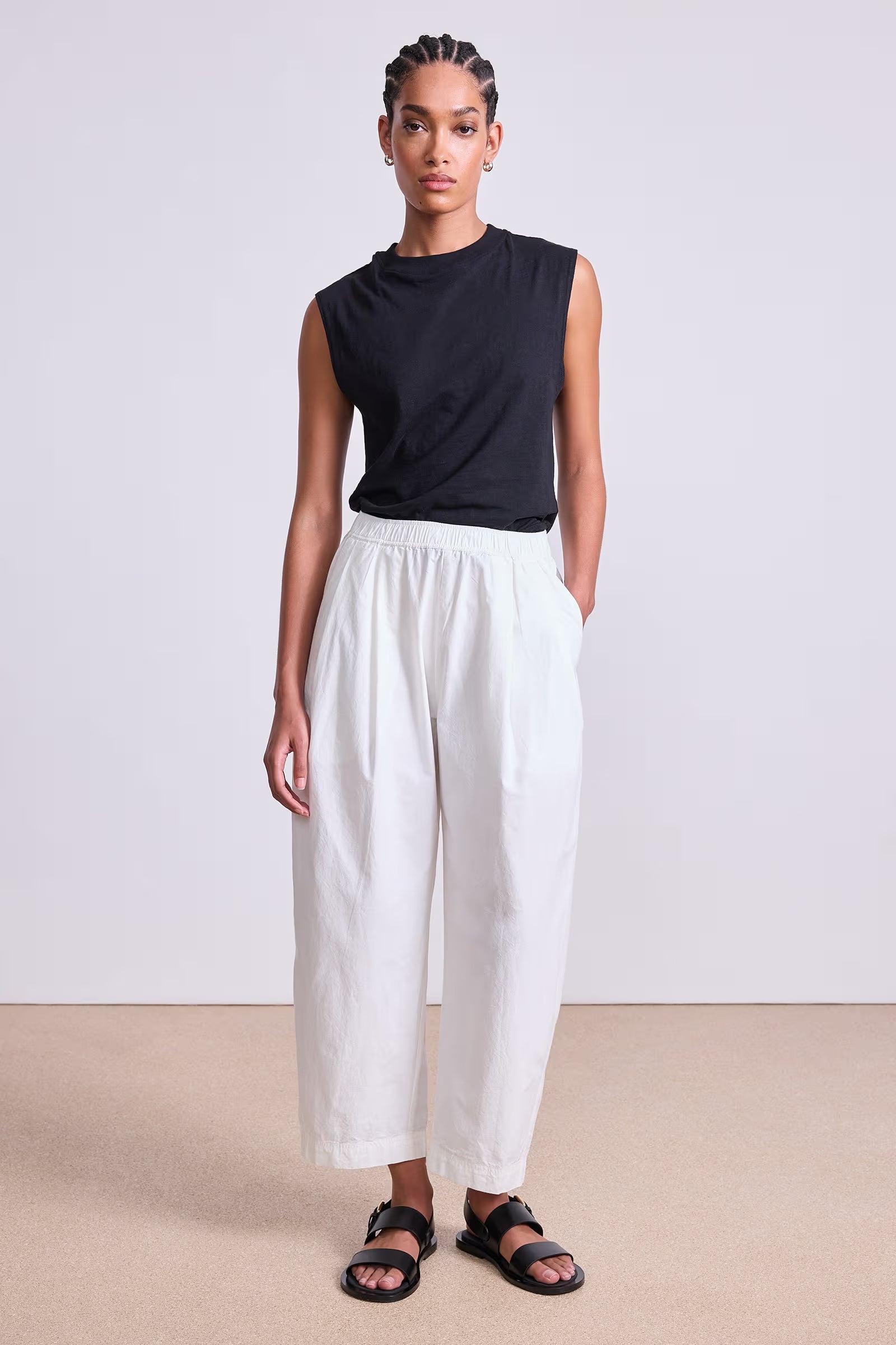Woman wearing a black sleeveless top and white pants against a plain background