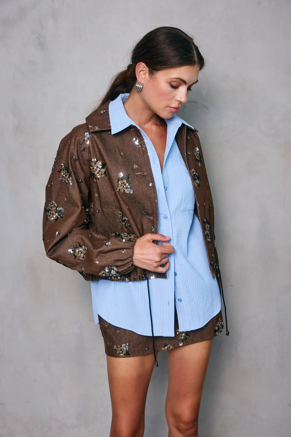 Woman wearing a brown embroidered jacket over a light blue shirt against a gray background, front