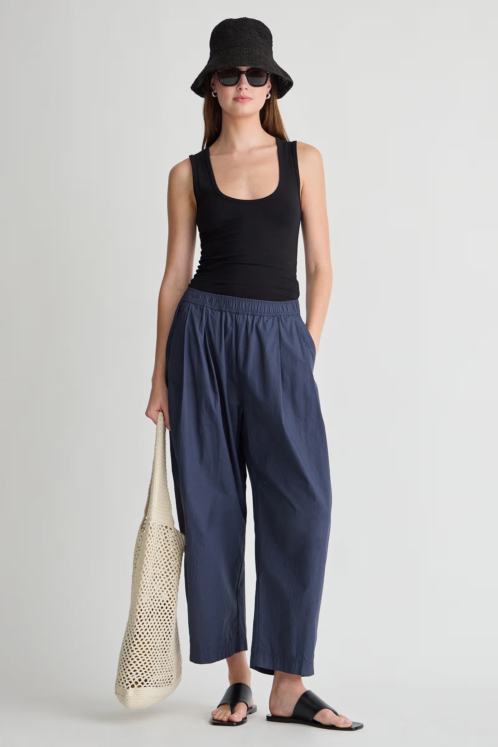 Woman wearing a black tank top and navy pants, holding a beige bag on a light gray background
