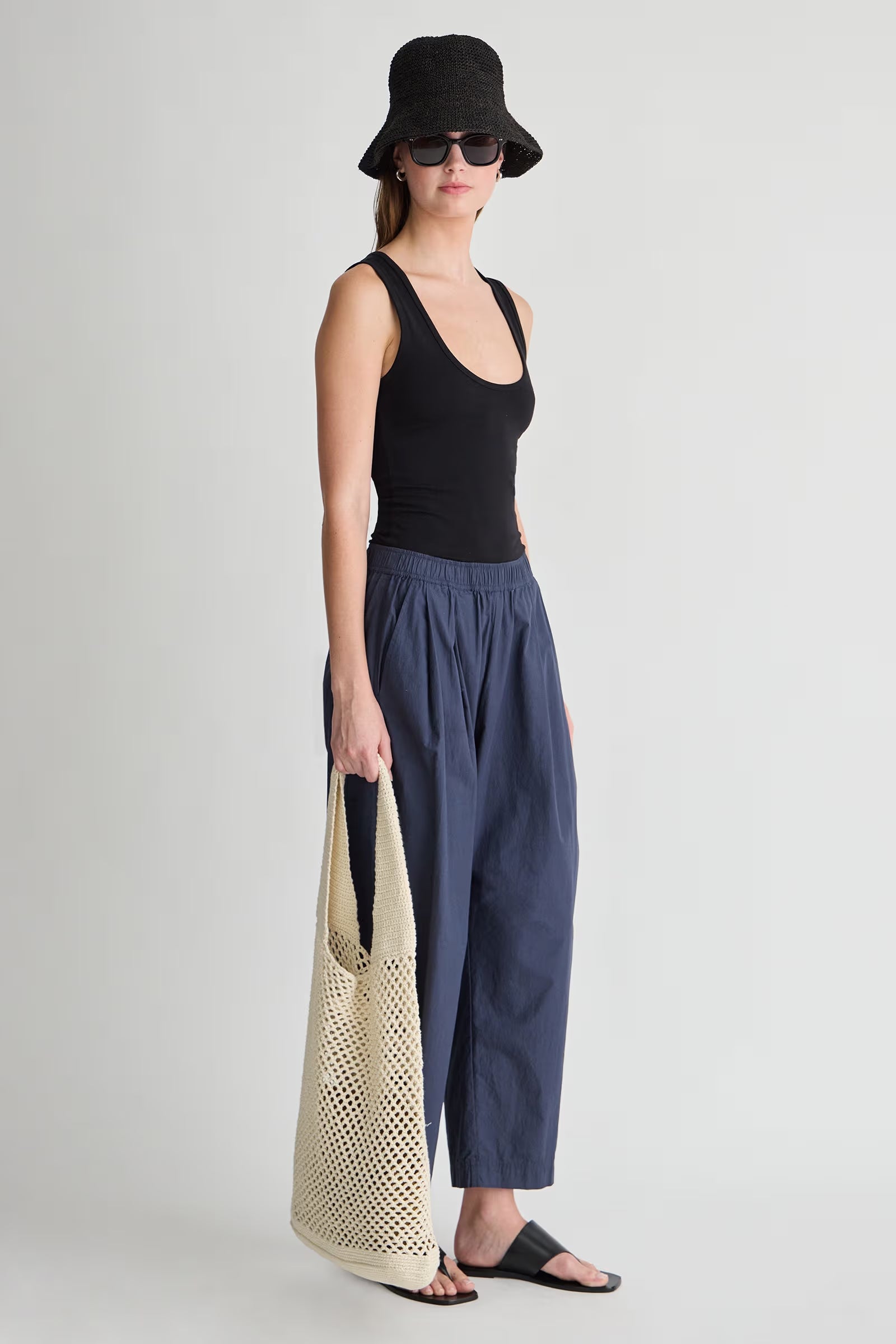 Woman wearing a black tank top and navy pants, holding a beige bag on a light gray background side