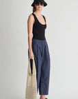 Woman wearing a black tank top and navy pants, holding a beige bag on a light gray background side