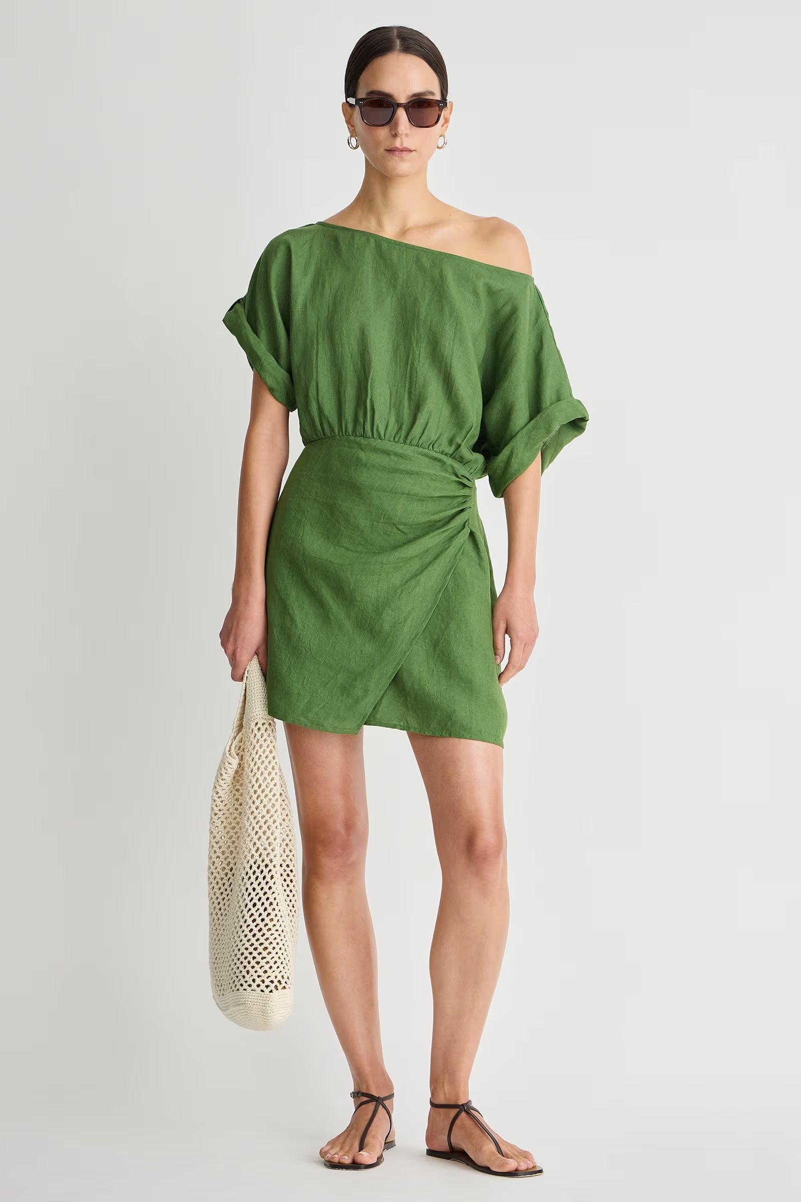 Woman wearing a green off-shoulder dress holding a beige woven bag on a white background
