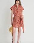 Woman wearing an orange patterned dress holding a yellow hat against a white background
