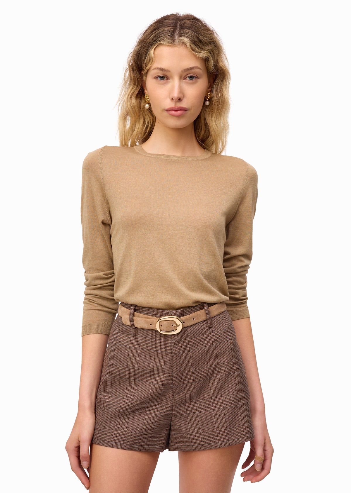 Woman wearing a beige long-sleeve top and brown shorts with a belt on a white background