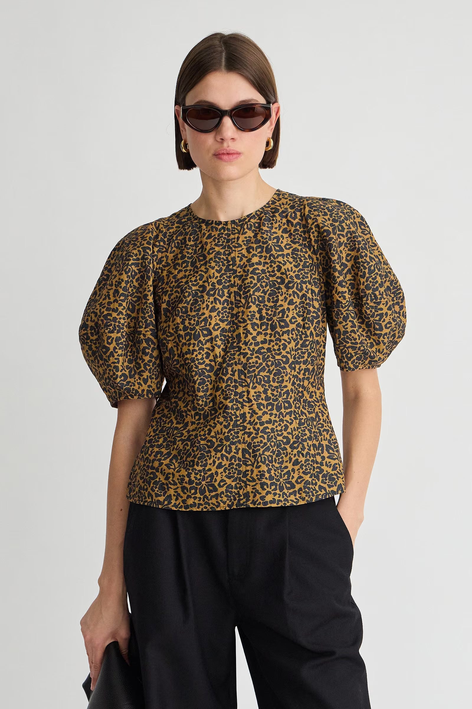 A model wearing a short-sleeved puff-sleeve blouse with a brown and black leopard print, paired with black trousers and sunglasses, against a plain light gray background.