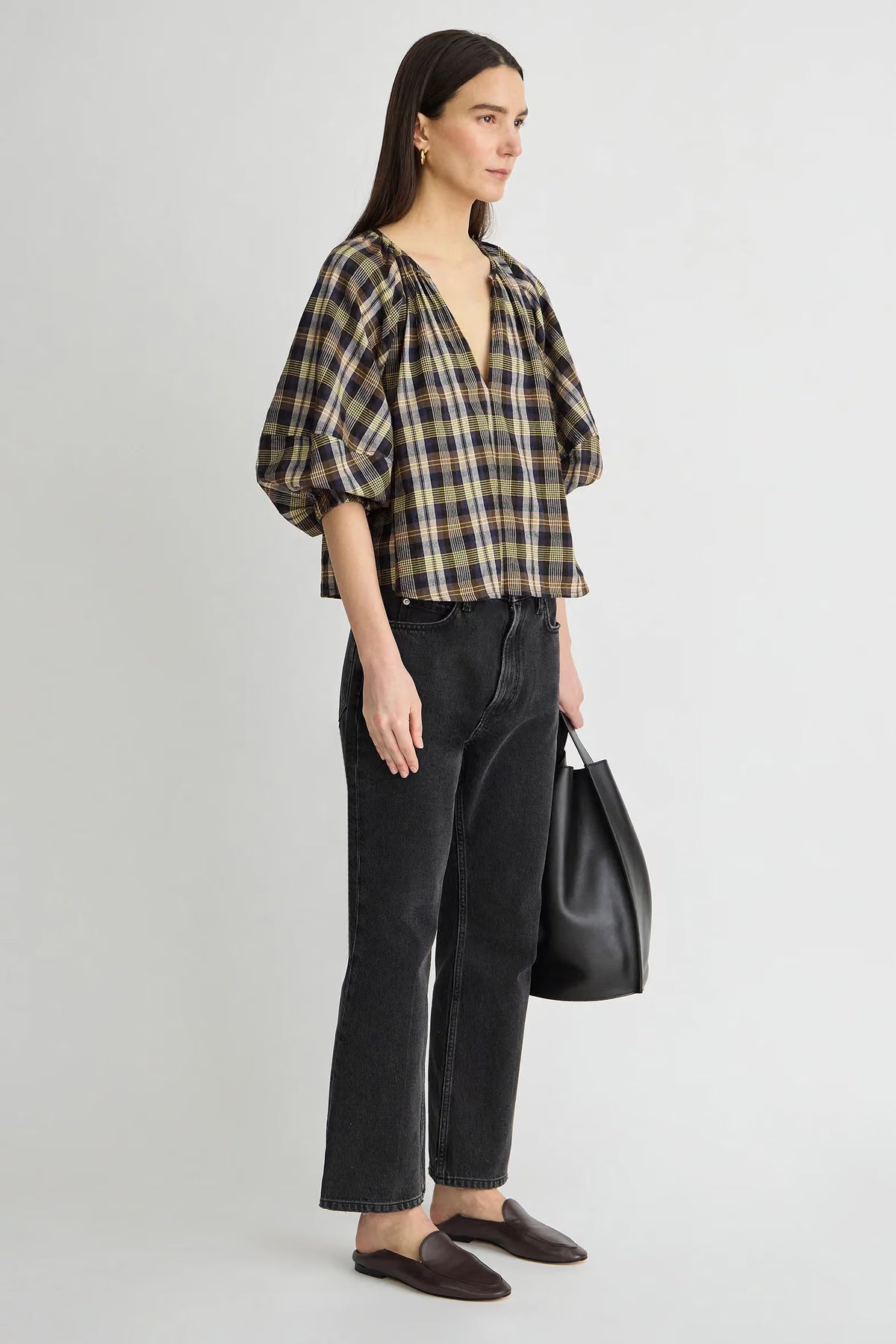 model wearing a plaid vneck short sleeve top with black jeans carrying a black bag on a grey background, side view