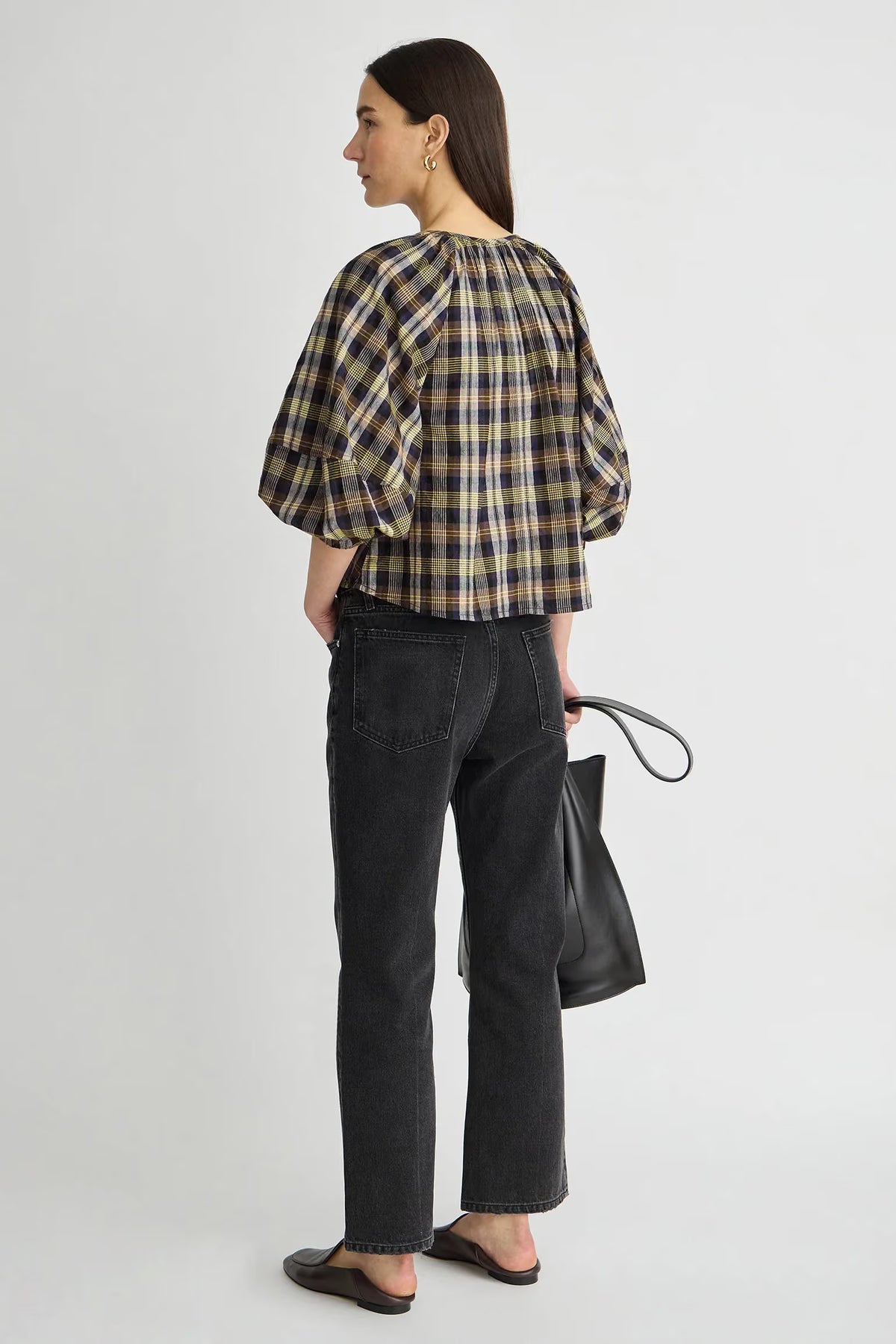 model wearing a plaid vneck short sleeve top with black jeans carrying a black bag on a grey background, back view 