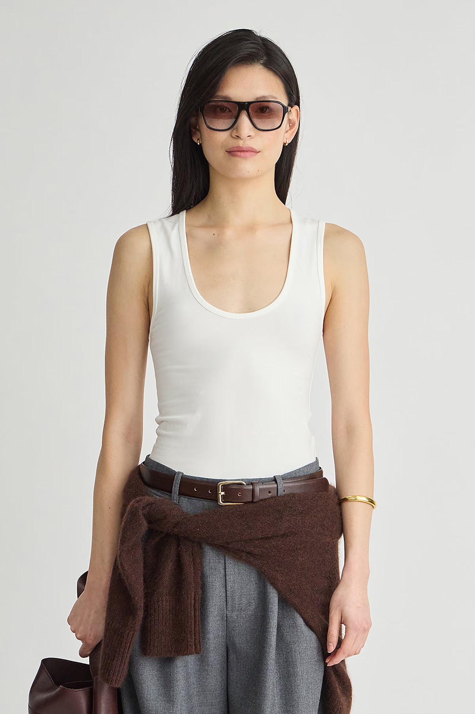 A model wearing a white ribbed scoop-neck tank top, gray wide-leg trousers, a brown belt, and a brown knit sweater tied around her waist, set against a plain light gray studio background.