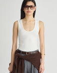 A model wearing a white ribbed scoop-neck tank top, gray wide-leg trousers, a brown belt, and a brown knit sweater tied around her waist, set against a plain light gray studio background.