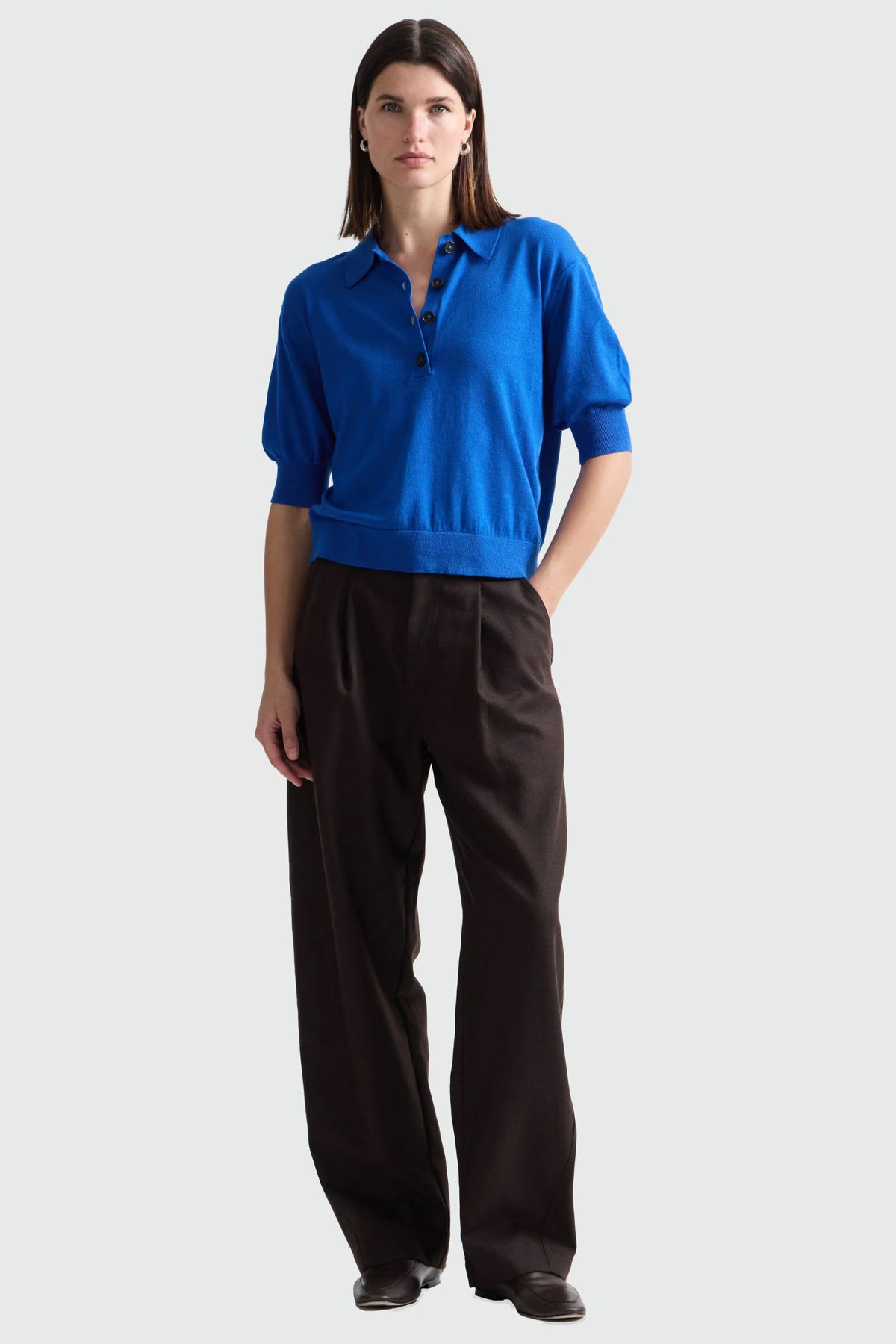 A woman modeling a bright blue short-sleeve knit polo shirt with dark buttons, paired with dark brown trousers, against a light gray studio background.