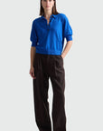 A woman modeling a bright blue short-sleeve knit polo shirt with dark buttons, paired with dark brown trousers, against a light gray studio background.