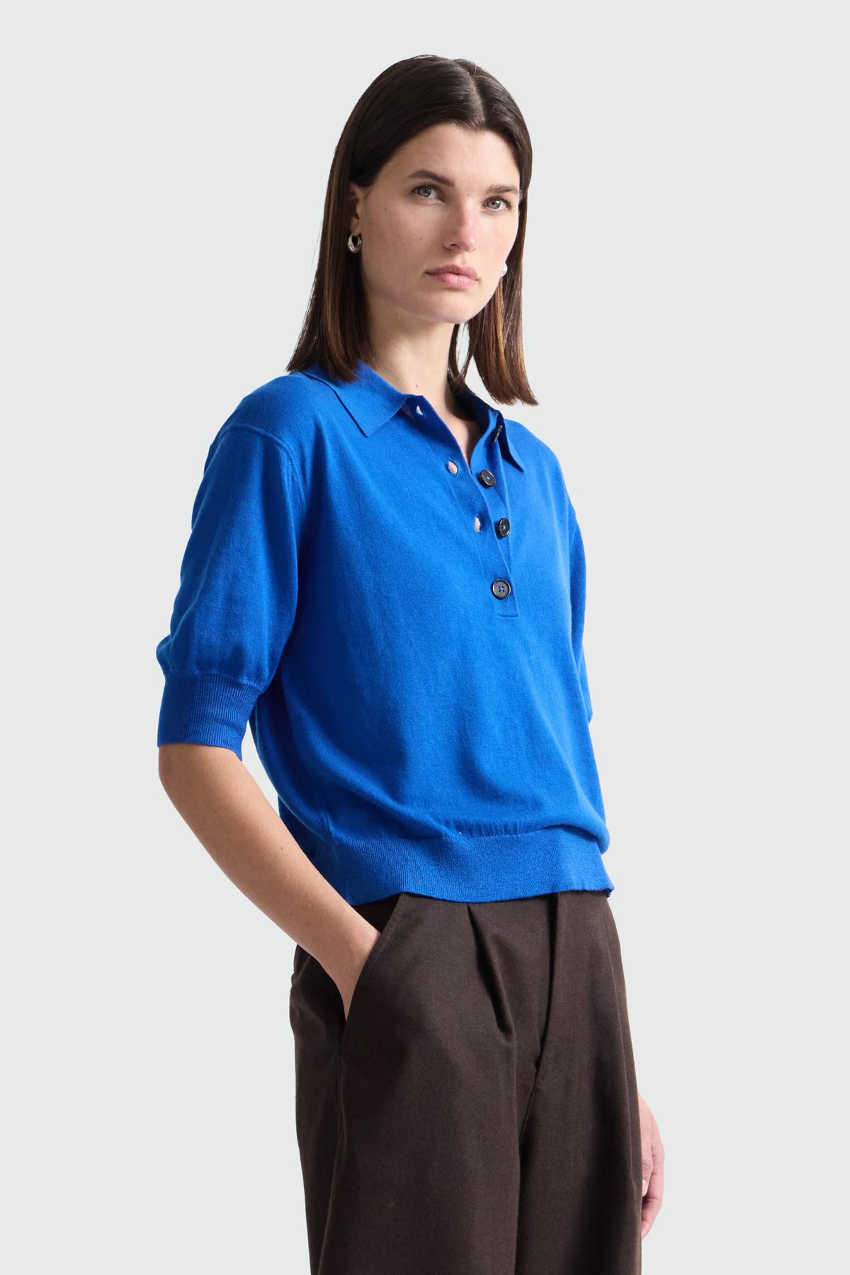 A woman modeling a bright blue short-sleeve knit polo shirt with dark buttons, paired with dark brown trousers, against a light gray studio background.