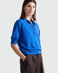 A woman modeling a bright blue short-sleeve knit polo shirt with dark buttons, paired with dark brown trousers, against a light gray studio background.