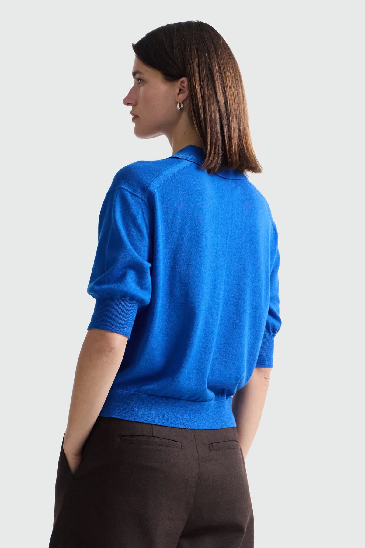 A woman shown from the back wearing a bright blue knit sweater with short puff sleeves and dark brown trousers, photographed against a clean white studio background.