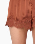 Close-up of a person wearing satin shorts in a warm rust or terracotta color, featuring a scalloped lace hem, shown against a plain white background.
