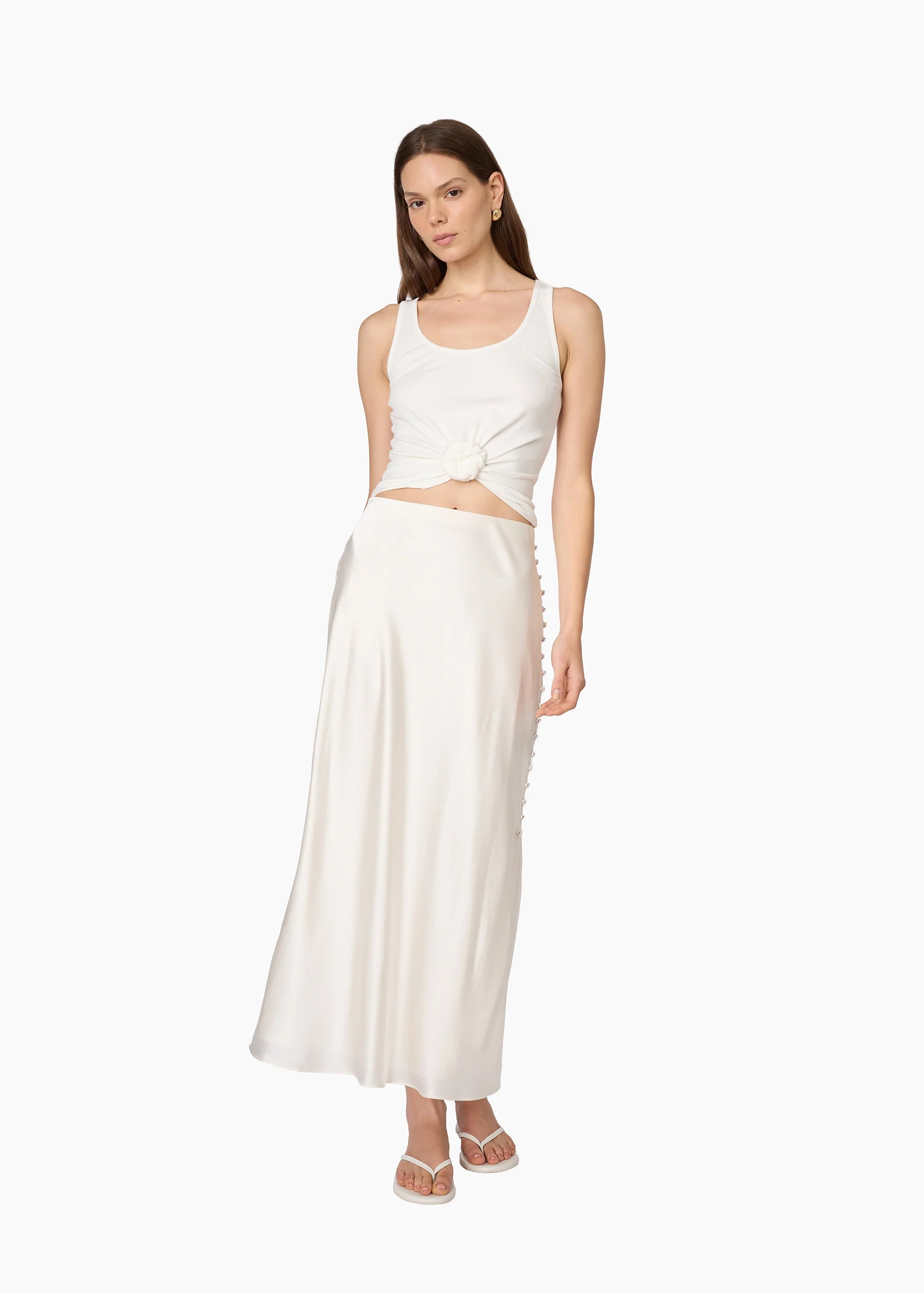 A woman modeling a two-piece white outfit consisting of a sleeveless crop top with a twisted detail at the waist and a matching long satin-like skirt, against a plain white background.