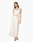 A woman modeling a two-piece white outfit consisting of a sleeveless crop top with a twisted detail at the waist and a matching long satin-like skirt, against a plain white background.