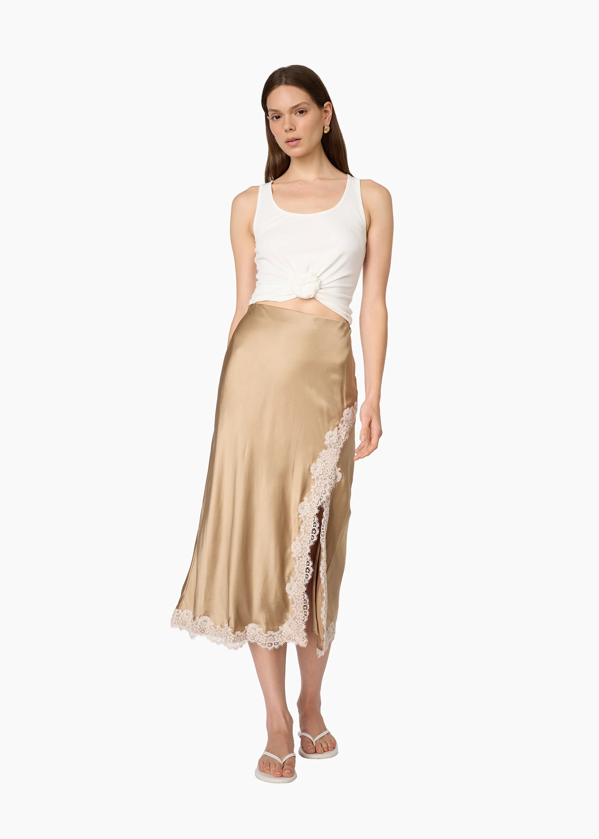 A woman modeling a champagne beige satin midi skirt with a lace side panel and hem, paired with a white crop top and white sandals, against a plain white background.