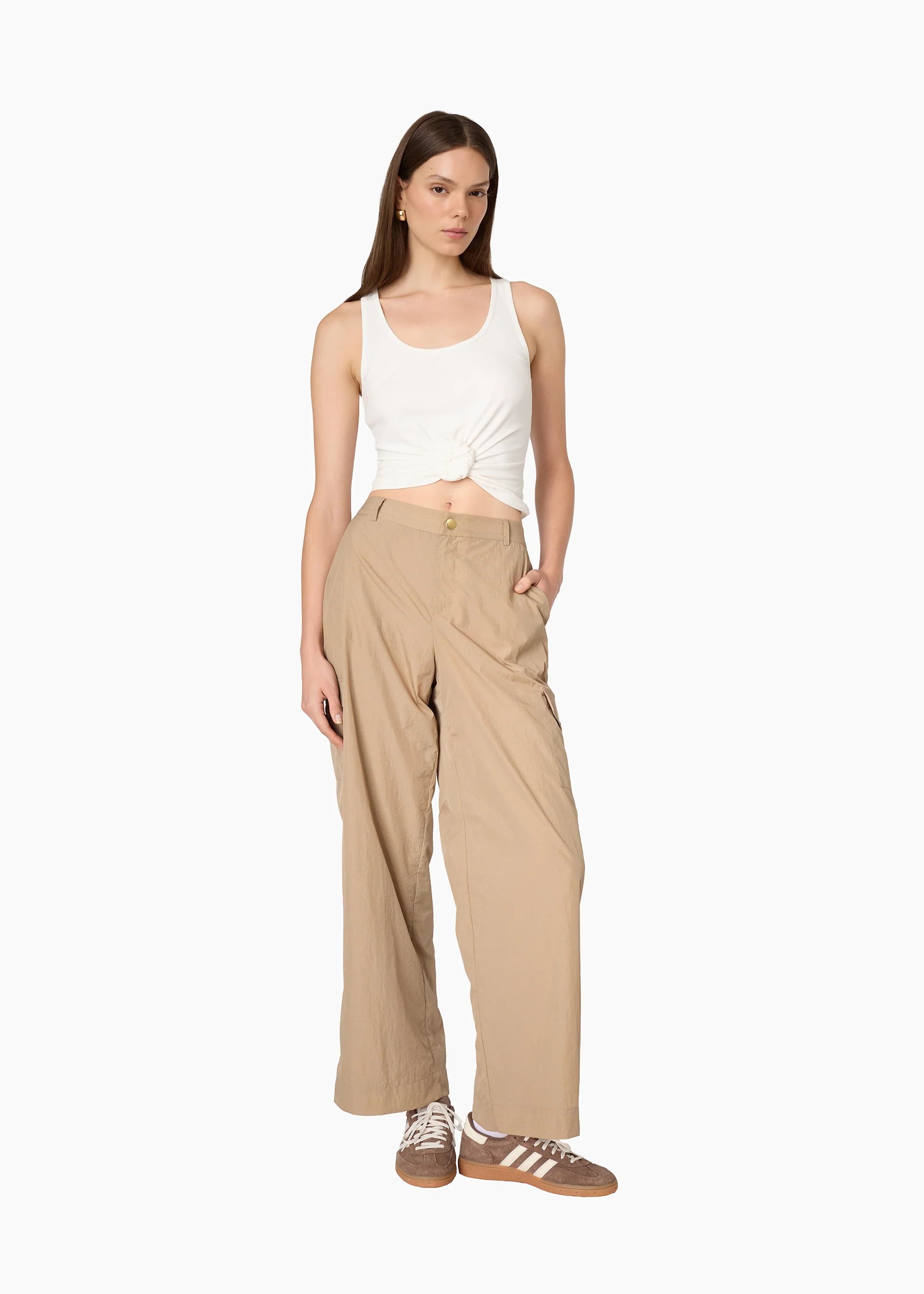 A model stands against a plain white background wearing beige wide-leg trousers, a white cropped tank top, and brown sneakers.