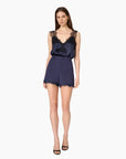 A woman modeling a navy blue satin romper with black lace shoulder panels and a lace hem, paired with black strappy heels, against a plain white background.