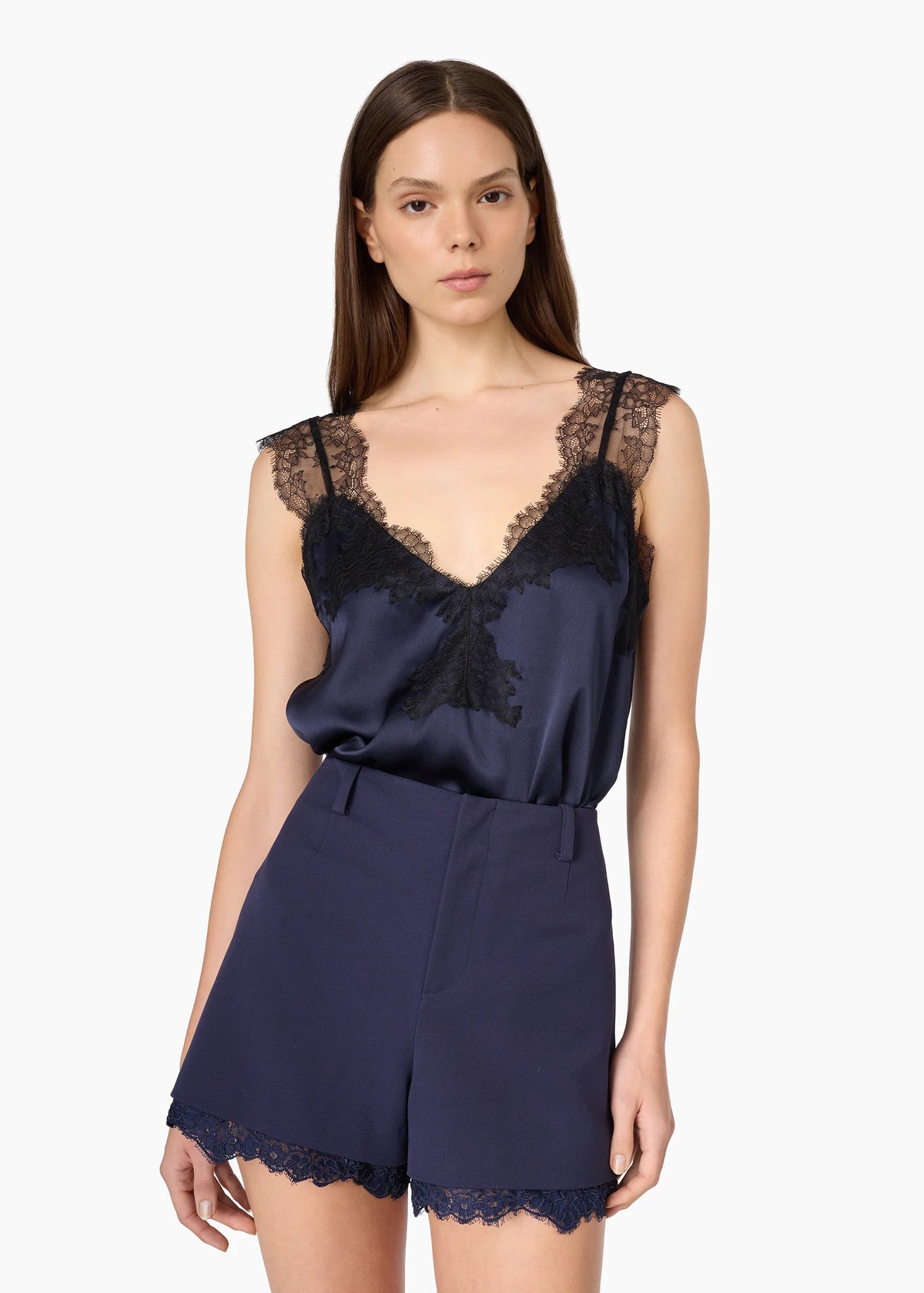 A woman modeling a navy blue satin camisole top with black lace trim and matching navy shorts with lace hem, against a plain white background.