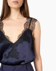 A close-up of a model wearing a navy blue satin camisole top with black lace trim along the V-neckline and shoulders, paired with navy trousers, against a plain white background.