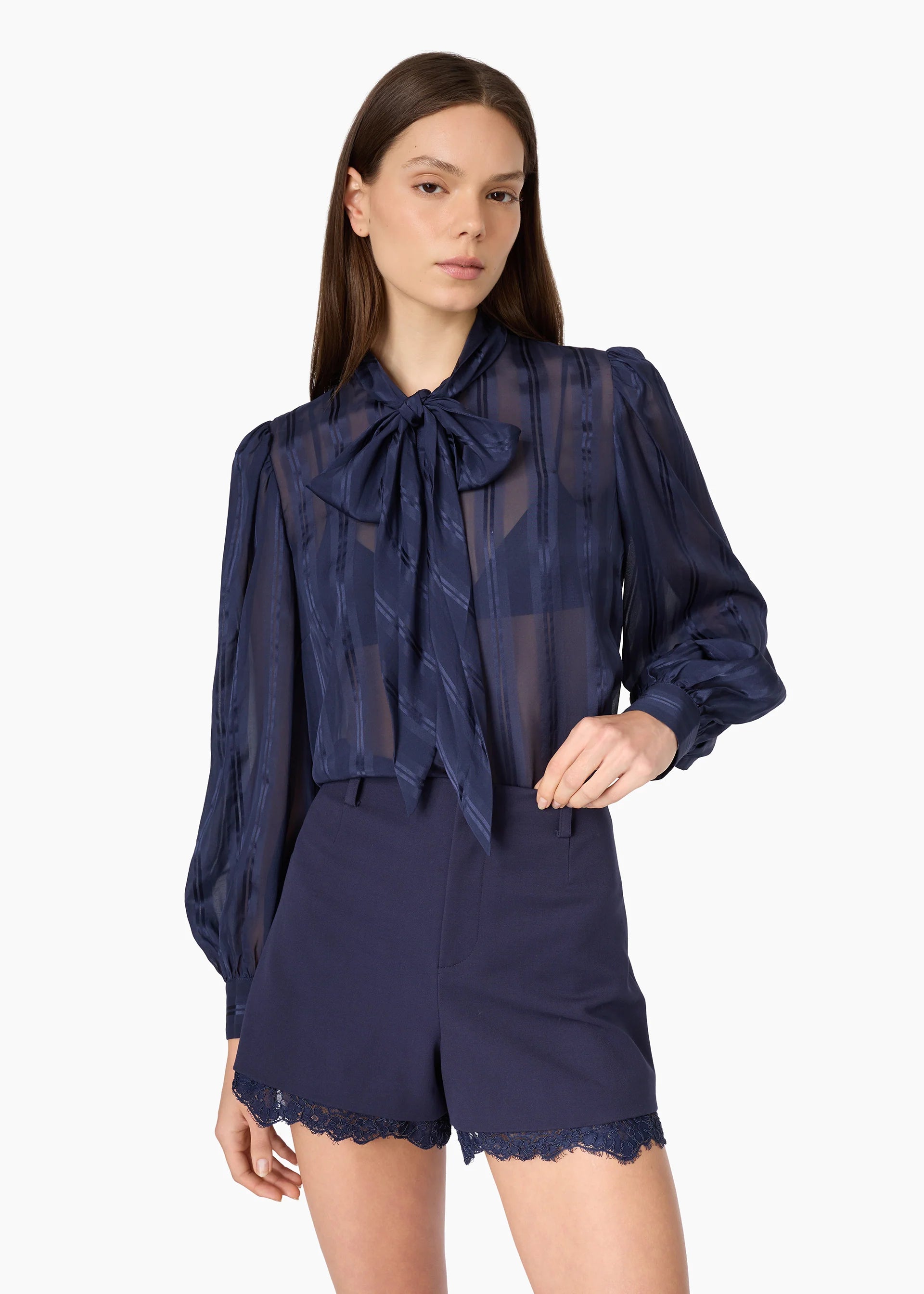 A model wearing a navy blue sheer long-sleeve blouse with a large bow tie-neck and matching navy blue shorts with a scalloped lace hem, set against a plain white studio background.