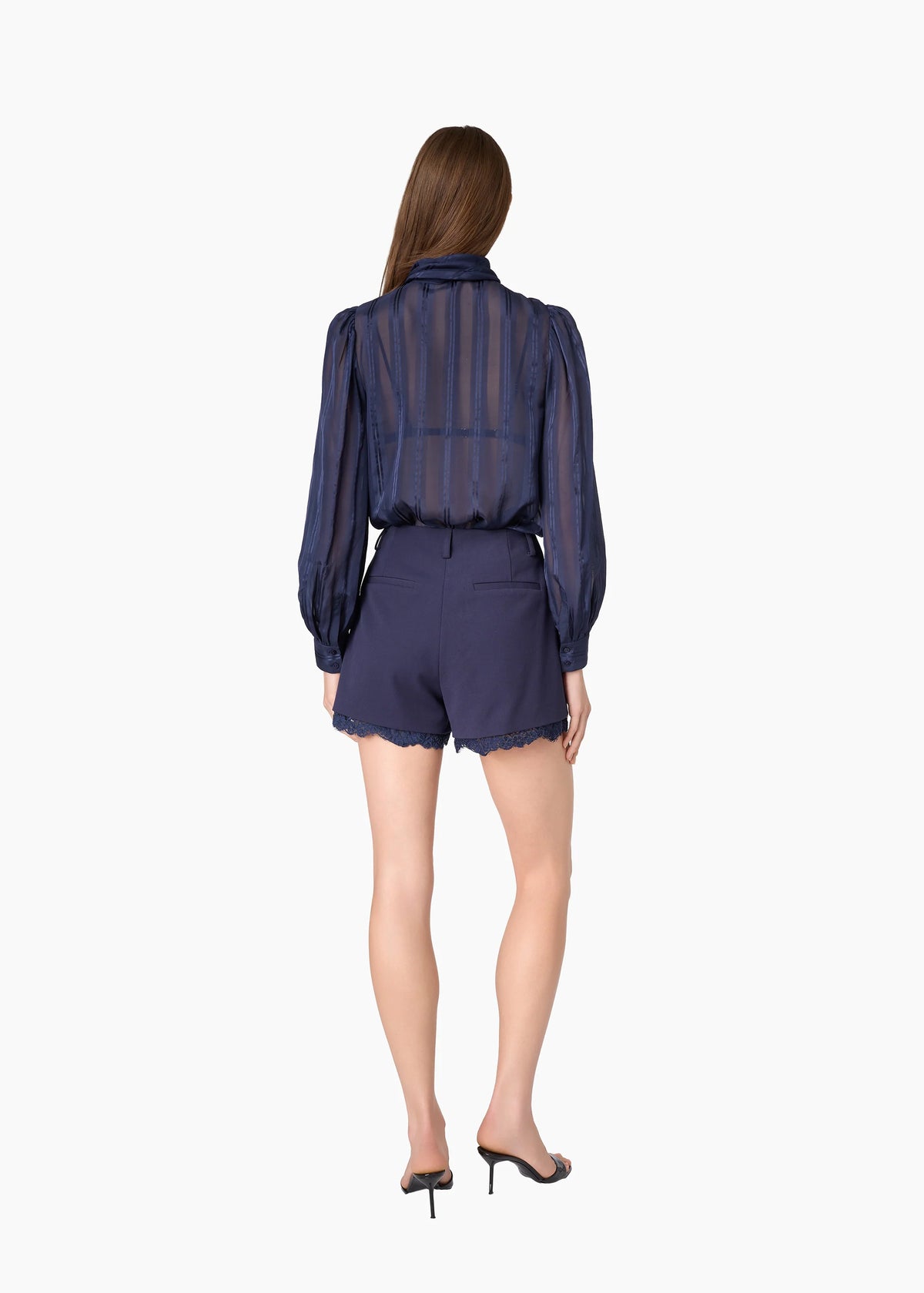 model wearing a navy sheer top with a tie neck with navy shorts that lace a small lace trim on a white background, back view
