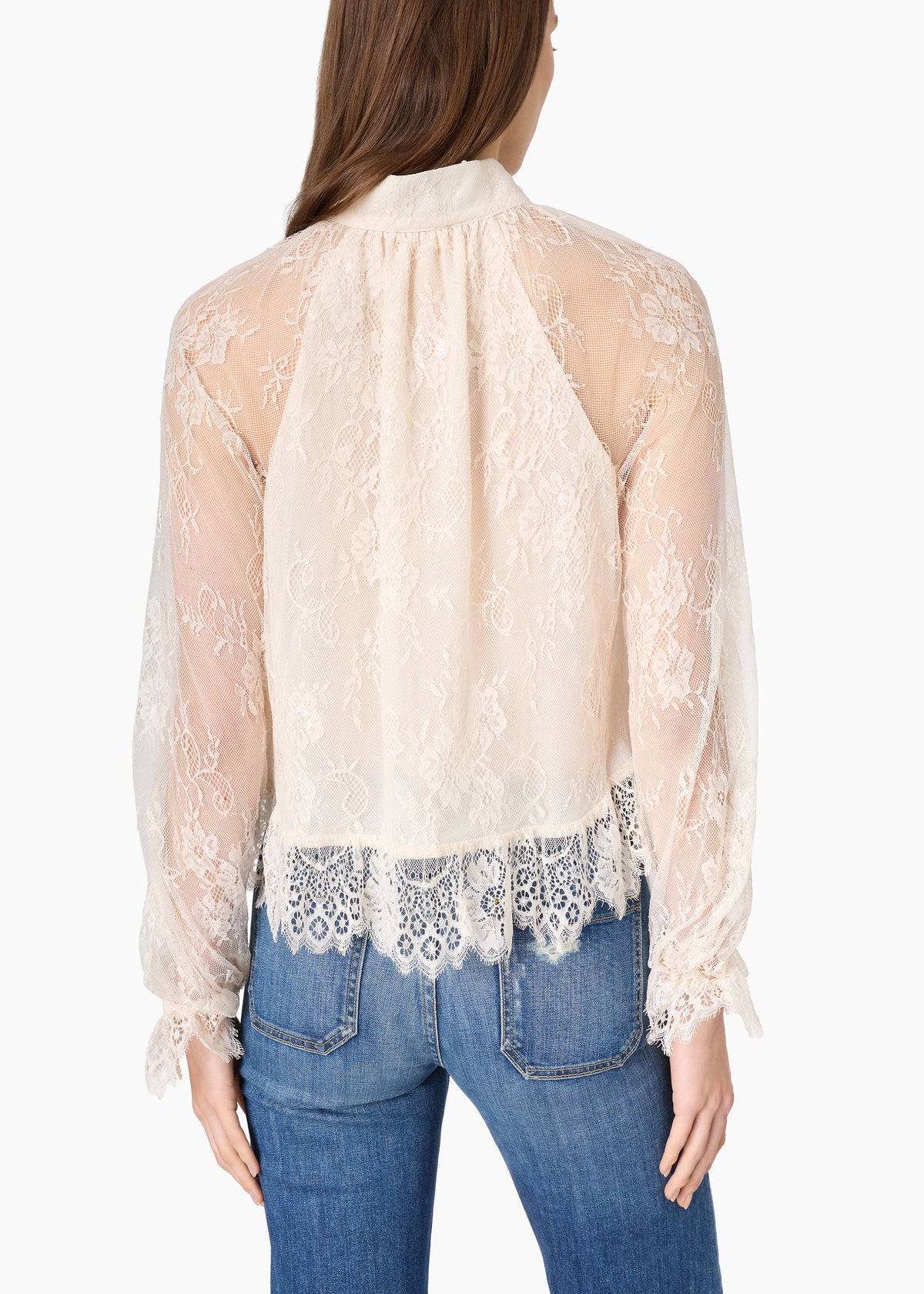 A woman viewed from the back wearing a sheer cream lace blouse with long sleeves and a gathered neckline, paired with blue jeans against a plain light background.
