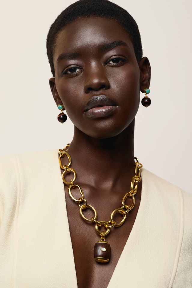 A close-up portrait of a model wearing a chunky gold chain necklace with a dark brown pendant, Umbria Earrings in Tigers Eye, and a cream-colored blazer against a light beige background.