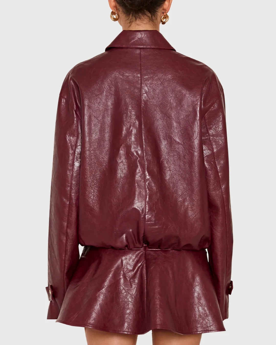 A person shown from the back wearing a glossy burgundy faux-leather jacket with a collar, long sleeves, and a gathered waist with a matching peplum-like flared section, against a light gray studio background.