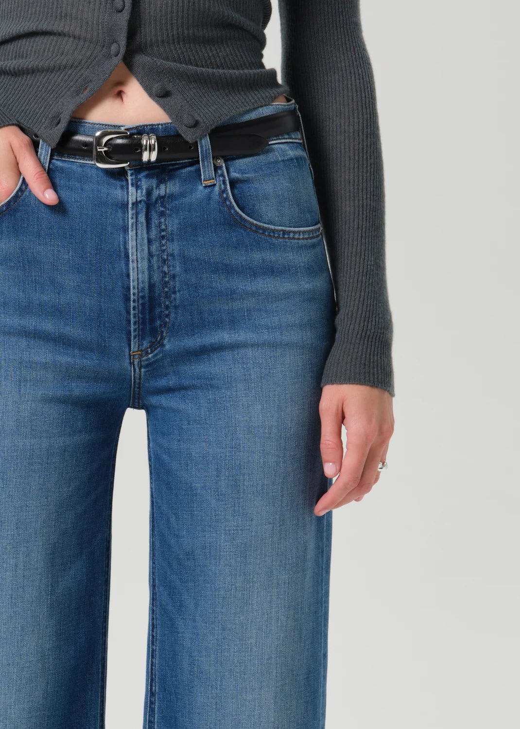 Close-up of a person wearing blue denim jeans with a black belt, paired with a dark gray ribbed top, against a light gray studio background.