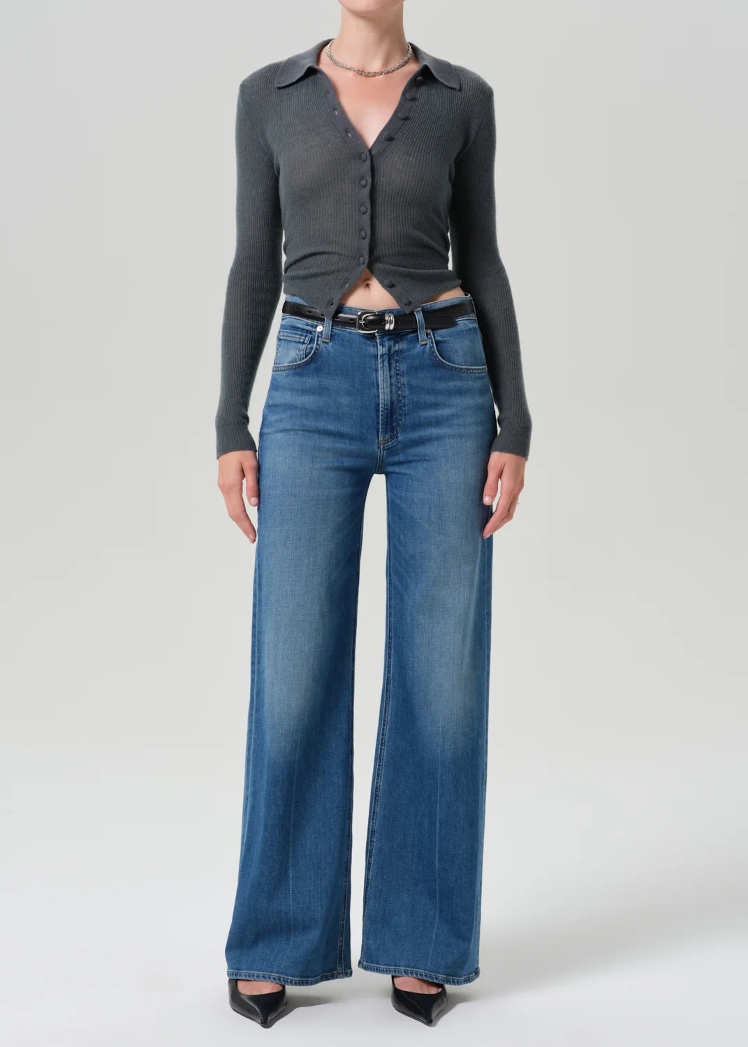 A model wearing a dark gray long-sleeve button-up shirt tucked into high-waisted, wide-leg blue jeans with a black belt, paired with black pointed-toe heels, against a light gray studio background.