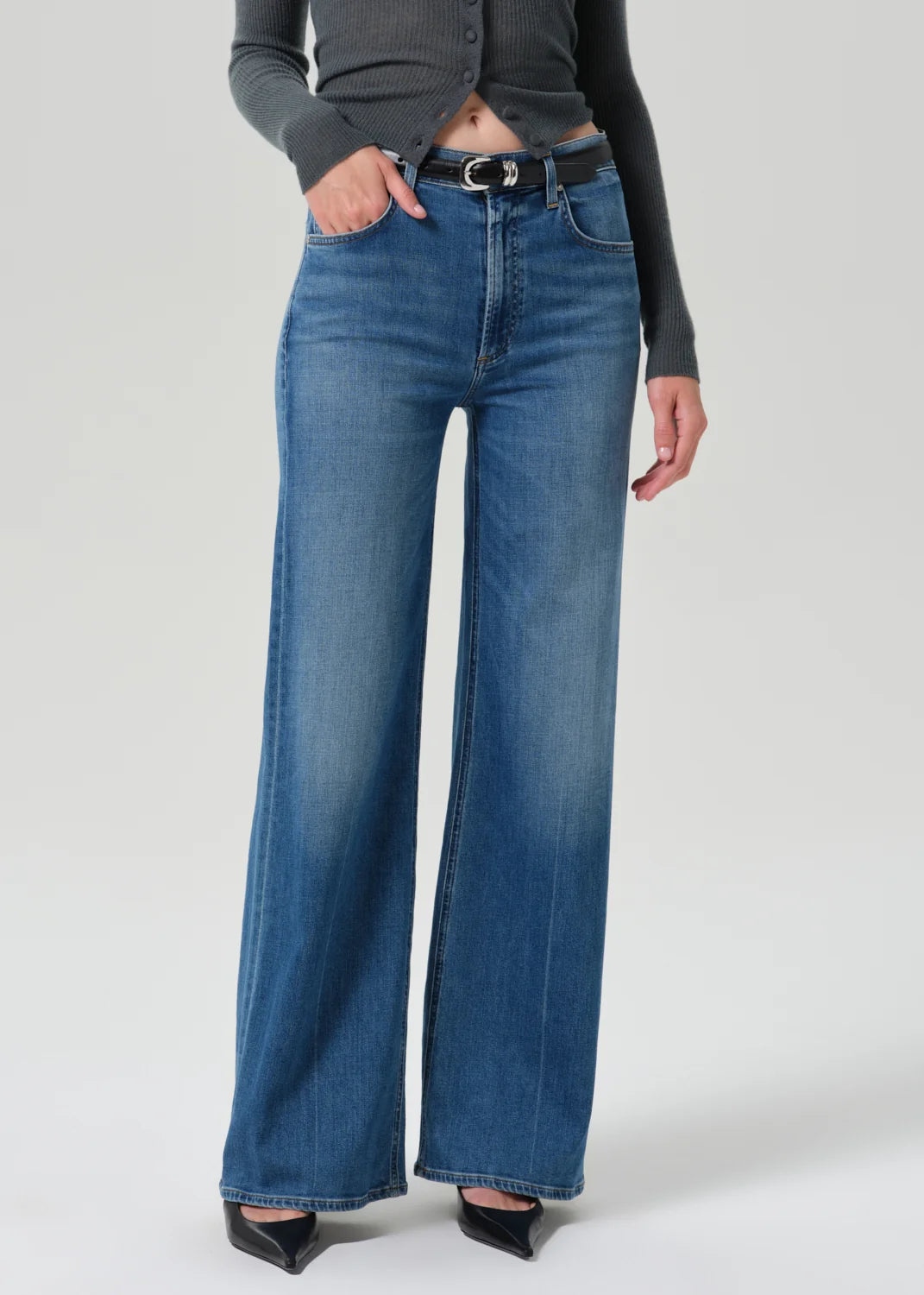 A model wearing a pair of high-waisted, wide-leg blue denim jeans with a black leather belt, styled with a dark gray long-sleeve top and black pointed-toe heels against a light gray studio background.