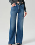A model wearing a pair of high-waisted, wide-leg blue denim jeans with a black leather belt, styled with a dark gray long-sleeve top and black pointed-toe heels against a light gray studio background.