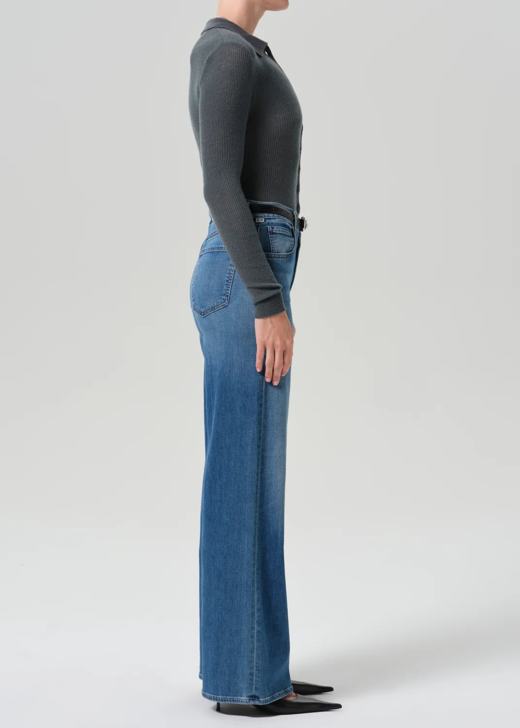 A side-profile studio shot of a model wearing a dark charcoal gray long-sleeve knit top and high-waisted, wide-leg blue denim jeans with a black belt.