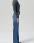 A side-profile studio shot of a model wearing a dark charcoal gray long-sleeve knit top and high-waisted, wide-leg blue denim jeans with a black belt.