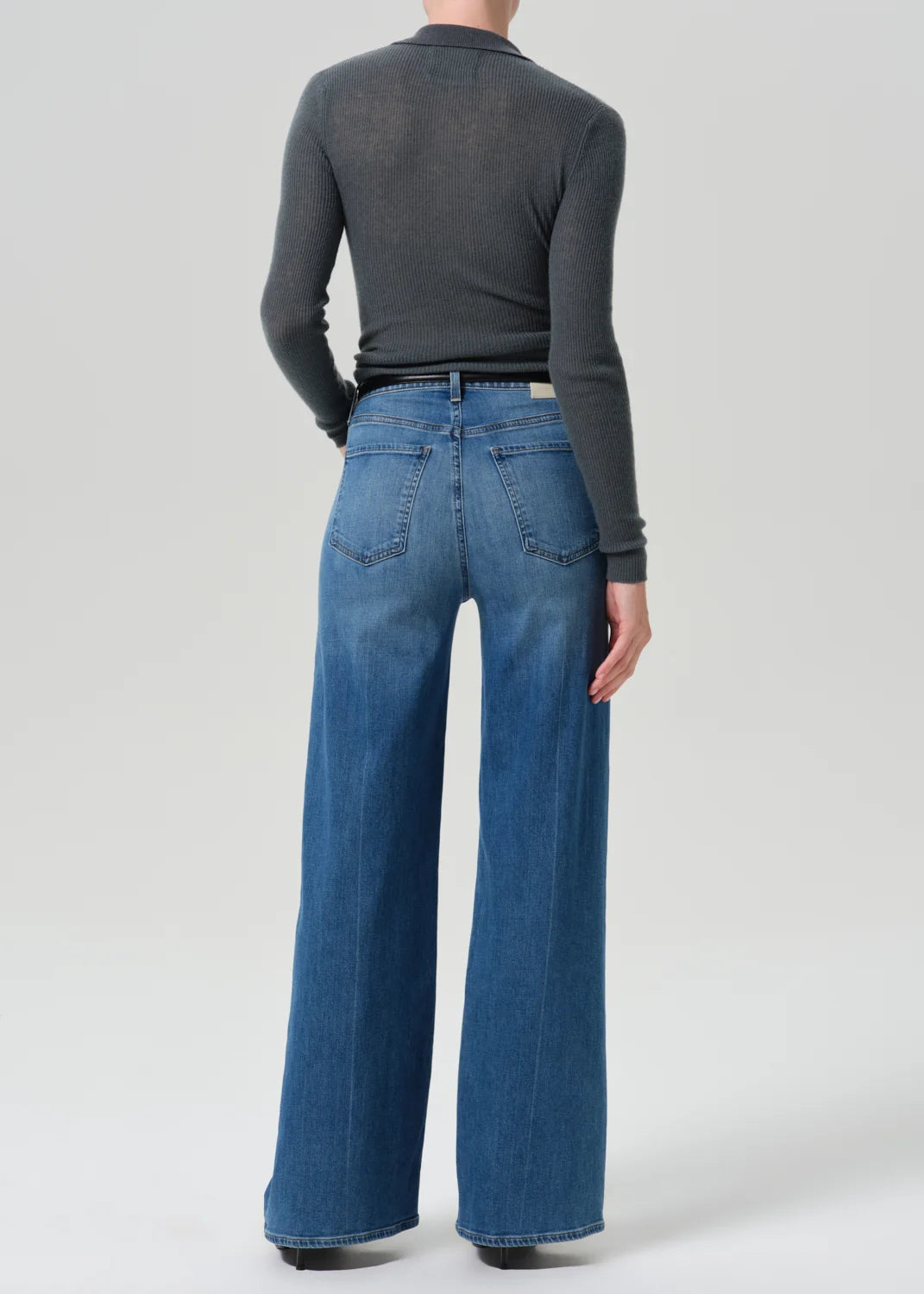 A model shown from the back wearing a dark gray long-sleeve mock neck top and high-waisted, wide-leg blue denim jeans against a light gray studio background.
