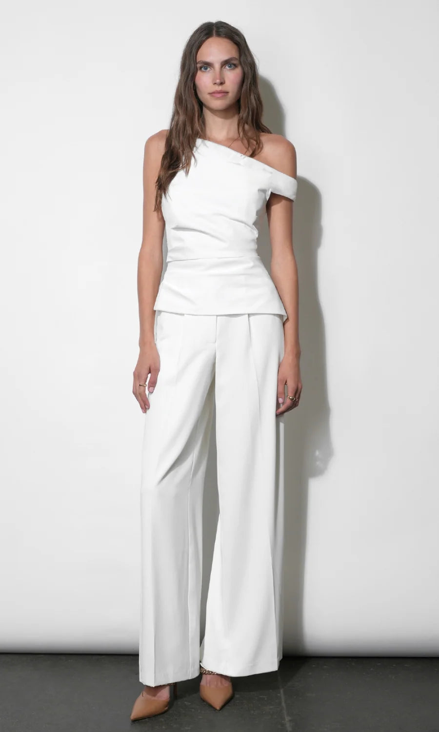 A woman modeling a white two-piece outfit consisting of an asymmetrical off-the-shoulder wrap top and matching wide-leg trousers, standing against a plain white wall.
