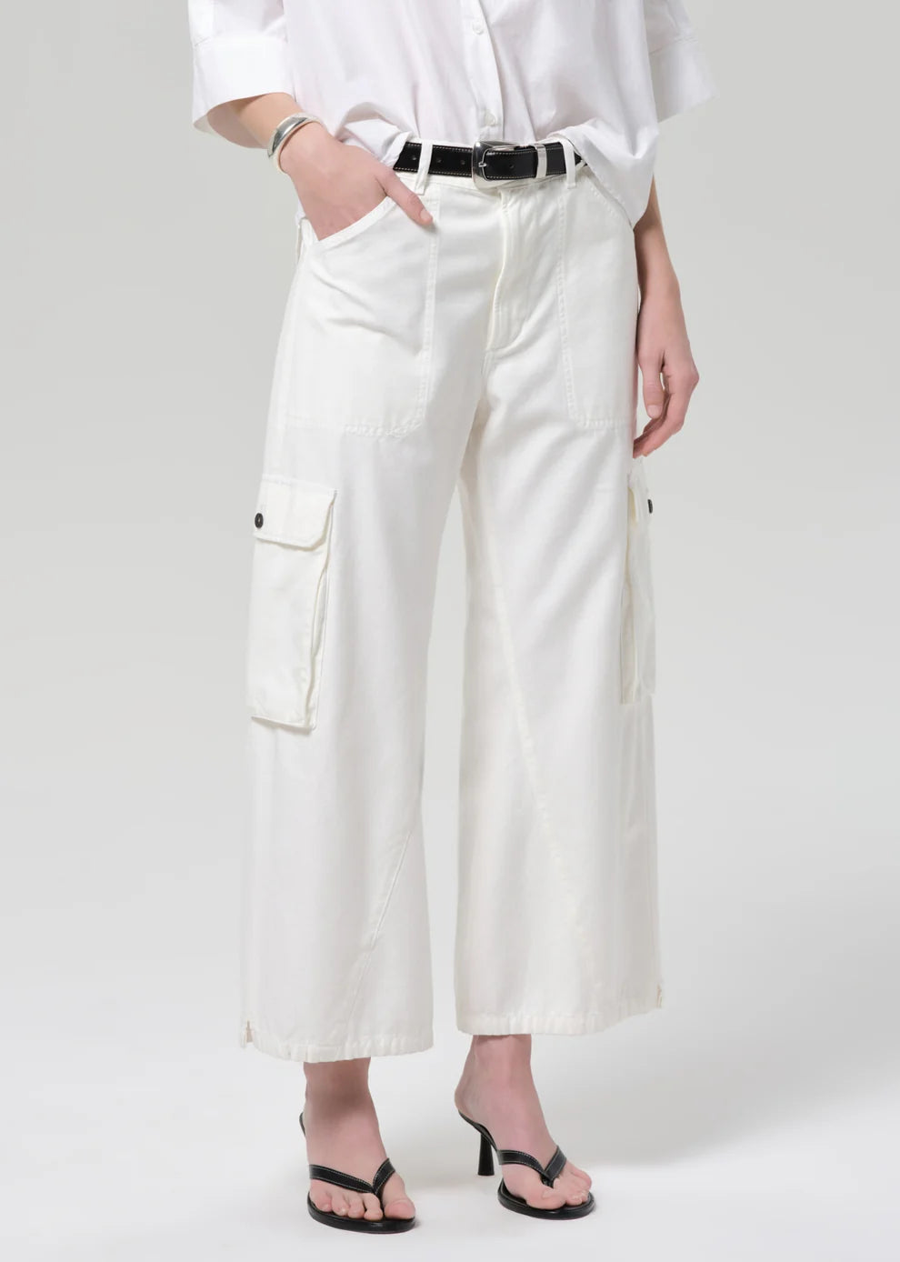 A model wearing white cropped cargo pants with a black belt, styled with a white shirt and black sandals against a light gray studio background.