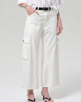 A model wearing white cropped cargo pants with a black belt, styled with a white shirt and black sandals against a light gray studio background.
