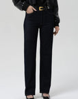 A model wearing high-waisted, straight-leg dark wash jeans with a gold-tone belt buckle, styled with a black leather jacket, black top, and black pointed-toe heels against a light gray studio background.