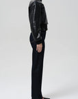 A model shown from the side wearing a black leather cropped jacket, black turtleneck, black wide-leg trousers, and black pointed-toe flats against a light gray studio background.