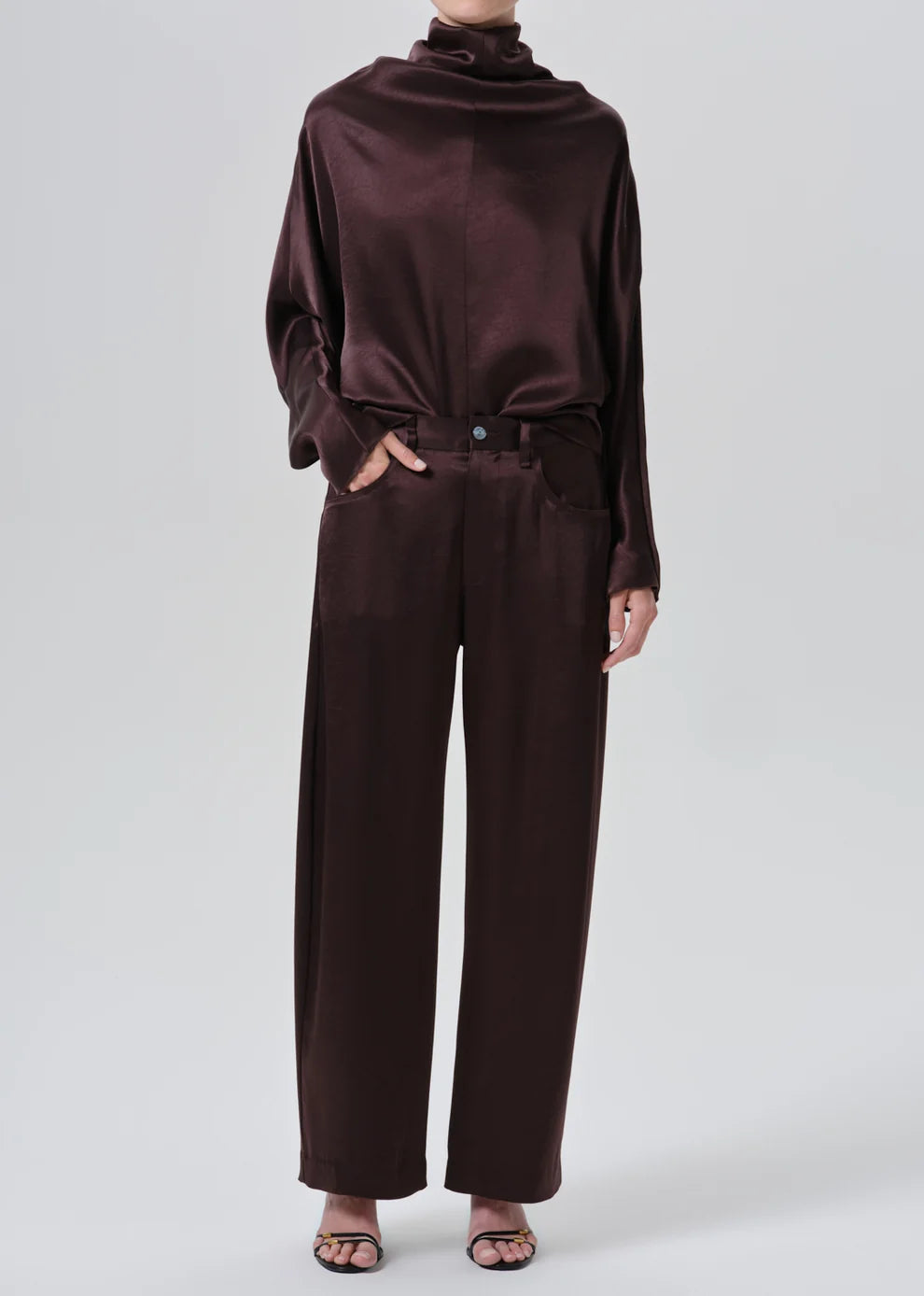 A model wearing a dark brown satin long-sleeve top with a cowl neck and matching wide-leg trousers, styled with black strappy heels against a light gray studio background.