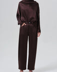 A model wearing a dark brown satin long-sleeve top with a cowl neck and matching wide-leg trousers, styled with black strappy heels against a light gray studio background.